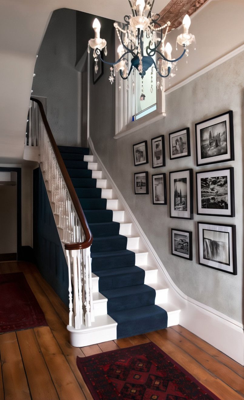A hallway with a blue-carpeted staircase, wooden floors, and framed black and white photos on a gray wall. A chandelier hangs above.