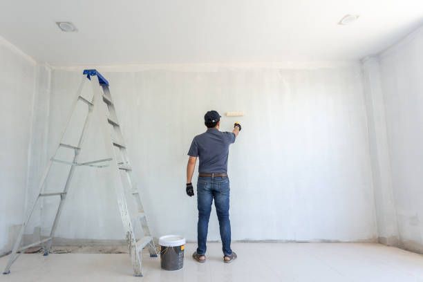 Person painting a white wall in an empty room, standing on the floor, using a paint roller. A ladder and paint bucket are also in the room.