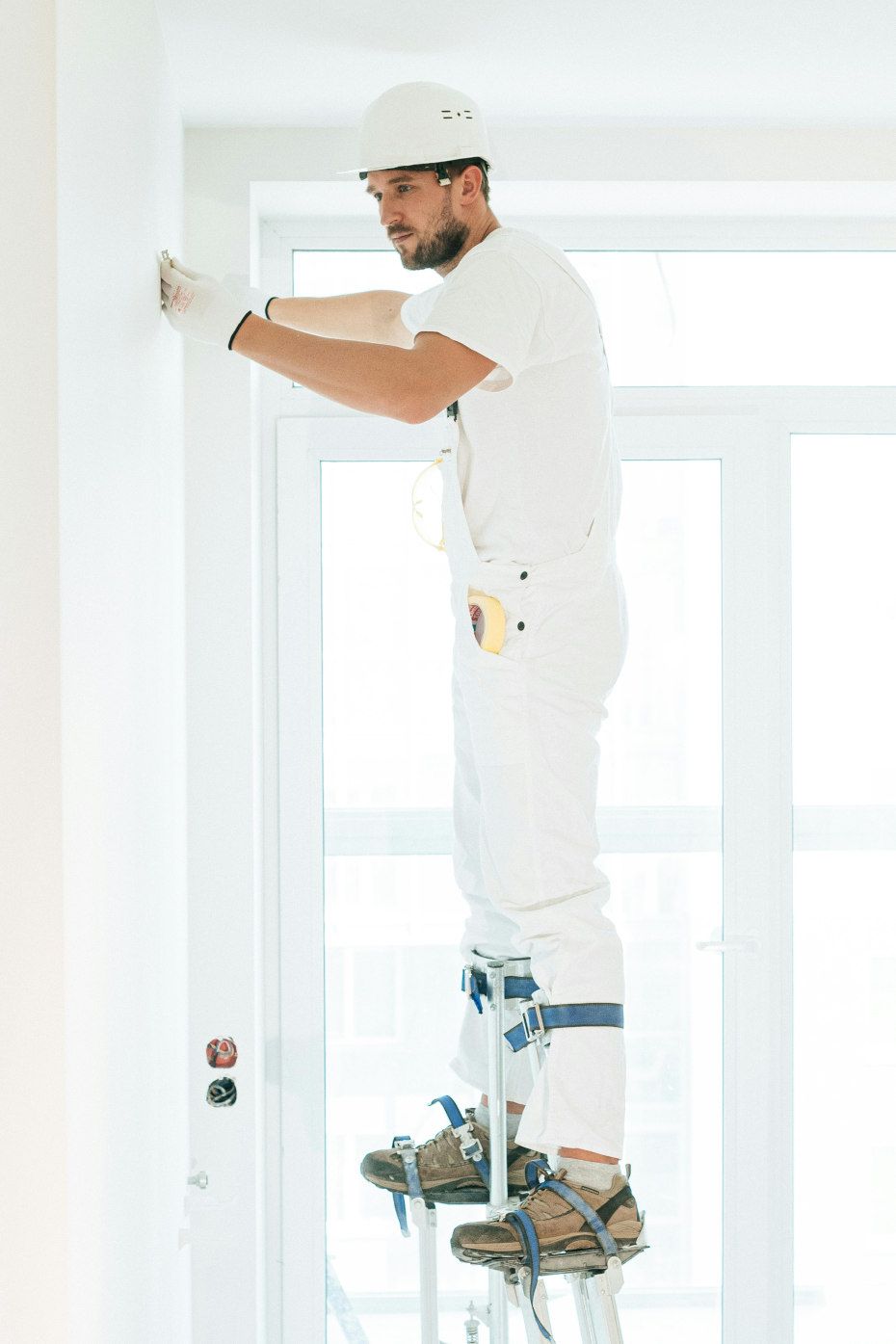 Painter in white overalls and hard hat on stilts reaching towards a wall, standing in front of a large window.