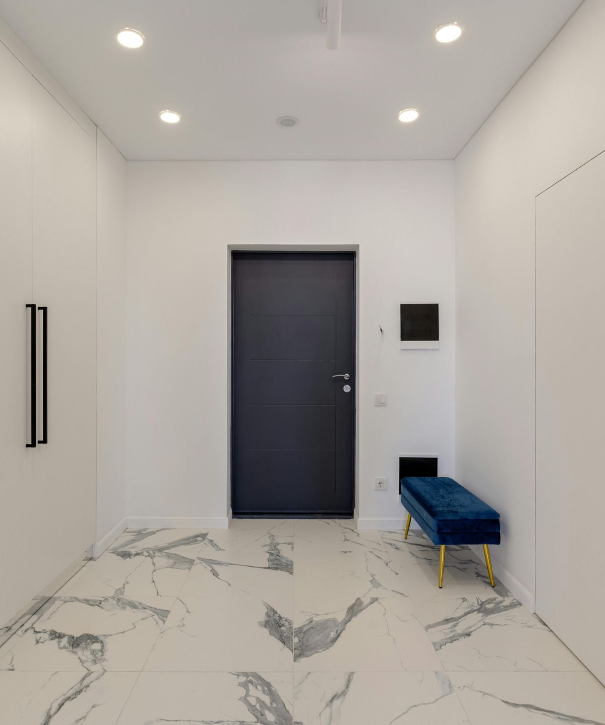 Hallway with white walls, marble-look tile floor, dark gray door, and blue velvet bench with gold legs.