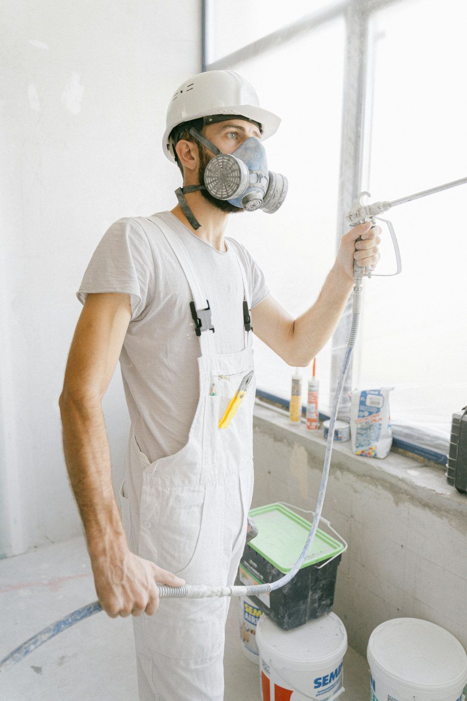 A construction worker in a respirator and overalls sprays sealant in a bright room.