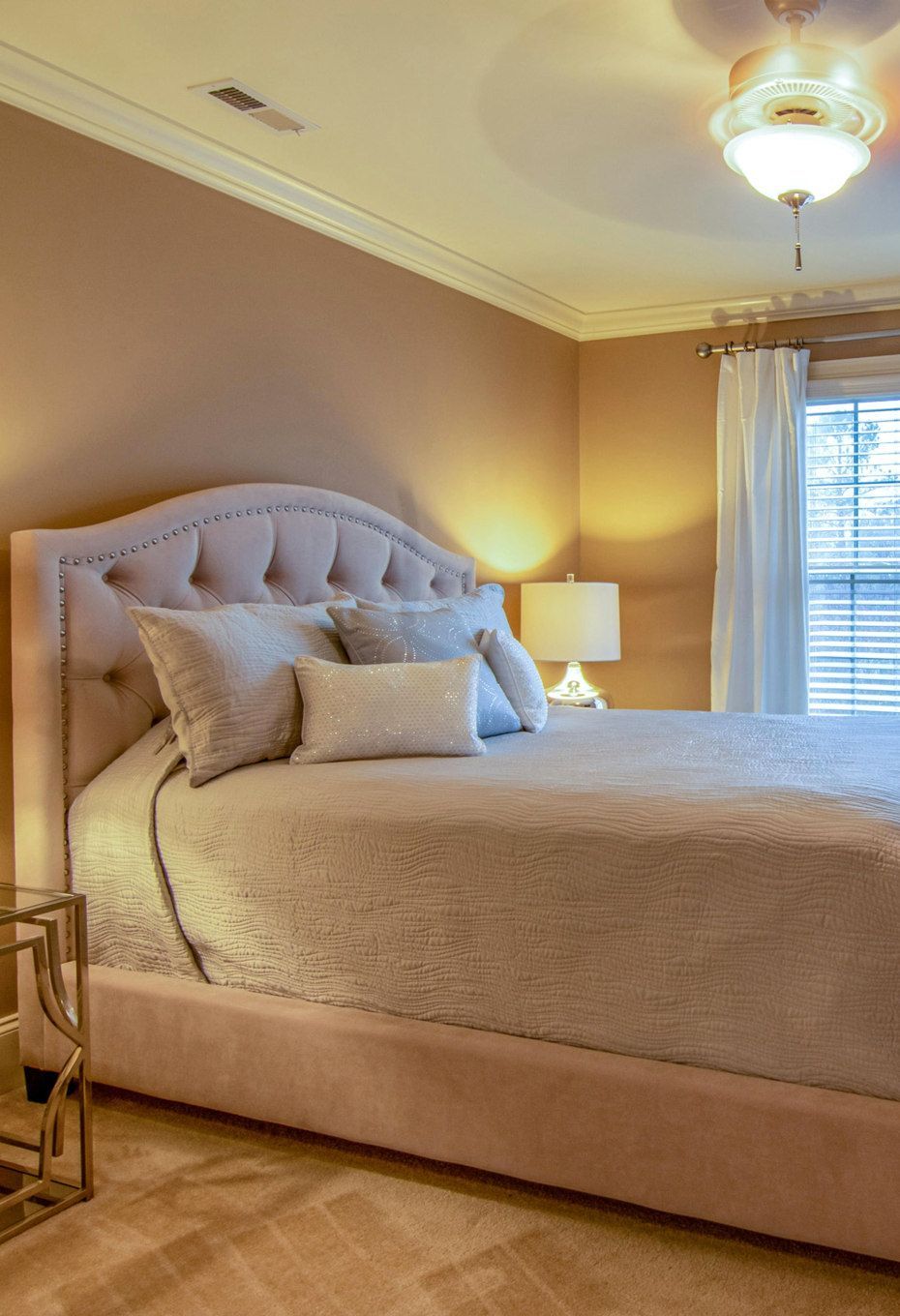 Bedroom with a large, upholstered bed in front of a window with blinds. Walls are peach, with soft lighting.
