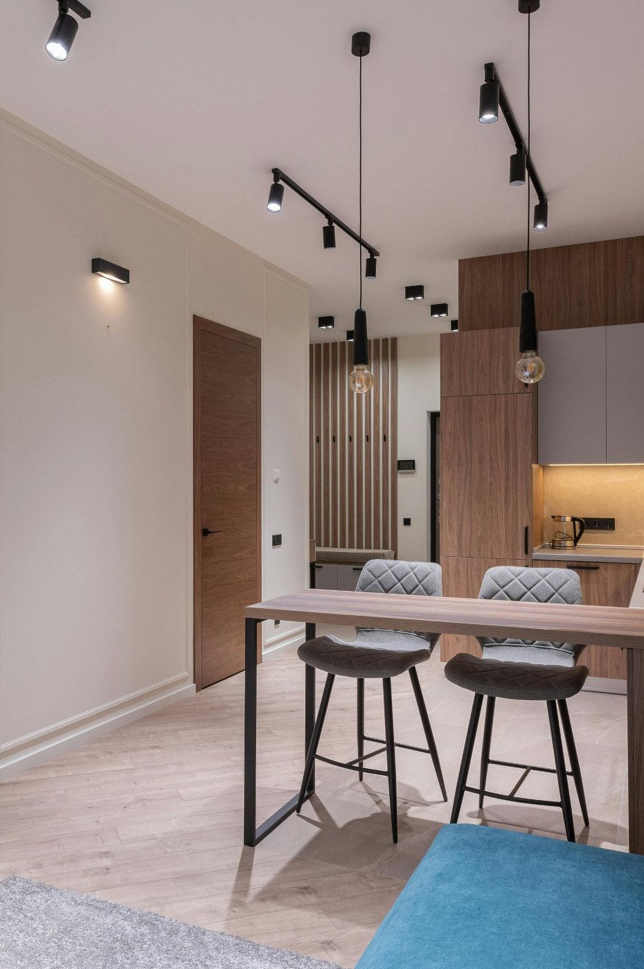 Modern kitchen and dining area with wooden accents, featuring a table with two grey stools under pendant lights.