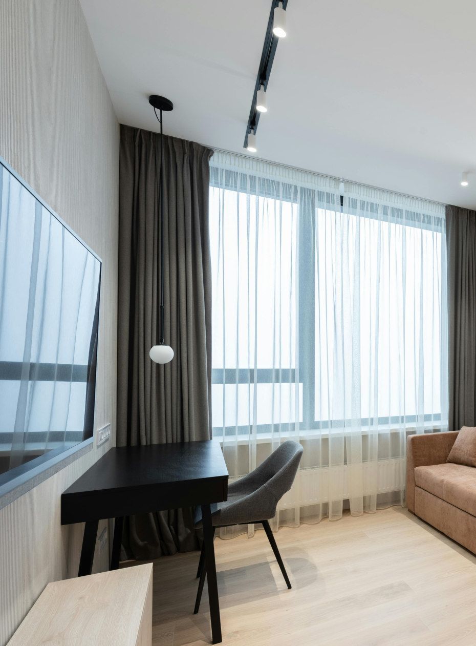 Desk and chair sit near a large window with sheer curtains and gray drapes. A TV hangs on the wall to the left.