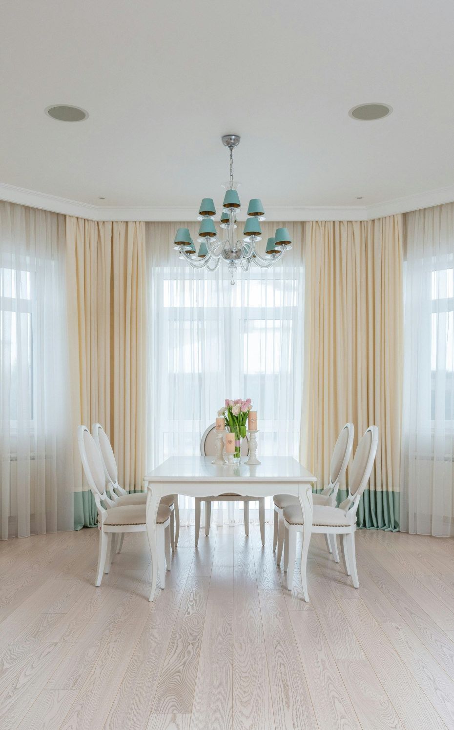 Elegant dining room with white table, chairs, and chandelier. Pale curtains frame a window, and the floor is a light wood.