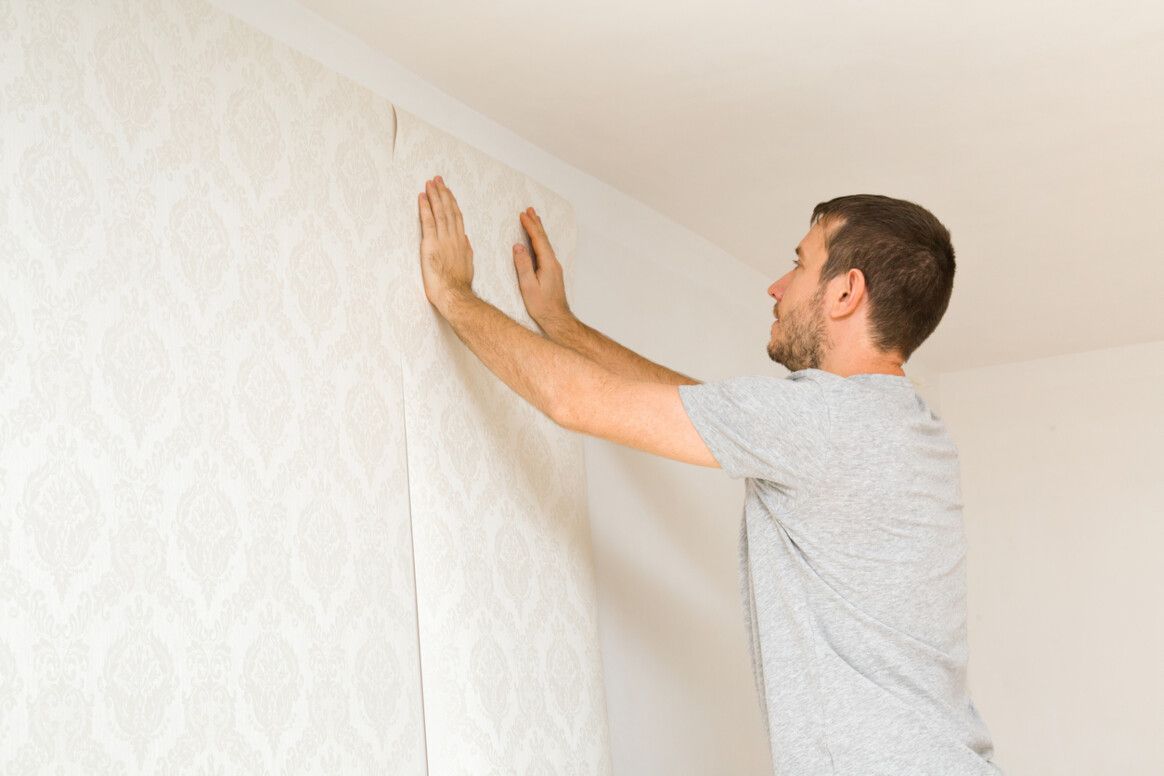 Man in gray shirt installing wallpaper, smoothing it against a white wall.