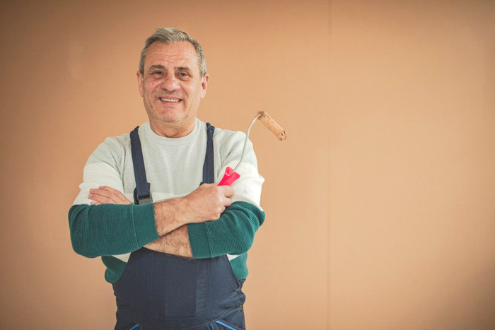 Man in blue overalls holding paint roller, smiling in front of a tan wall.