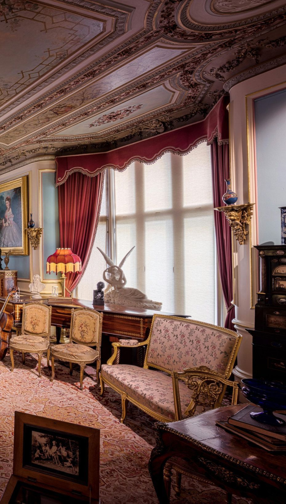 Ornate room with red drapes, gold furniture, and detailed ceiling. A window in center.