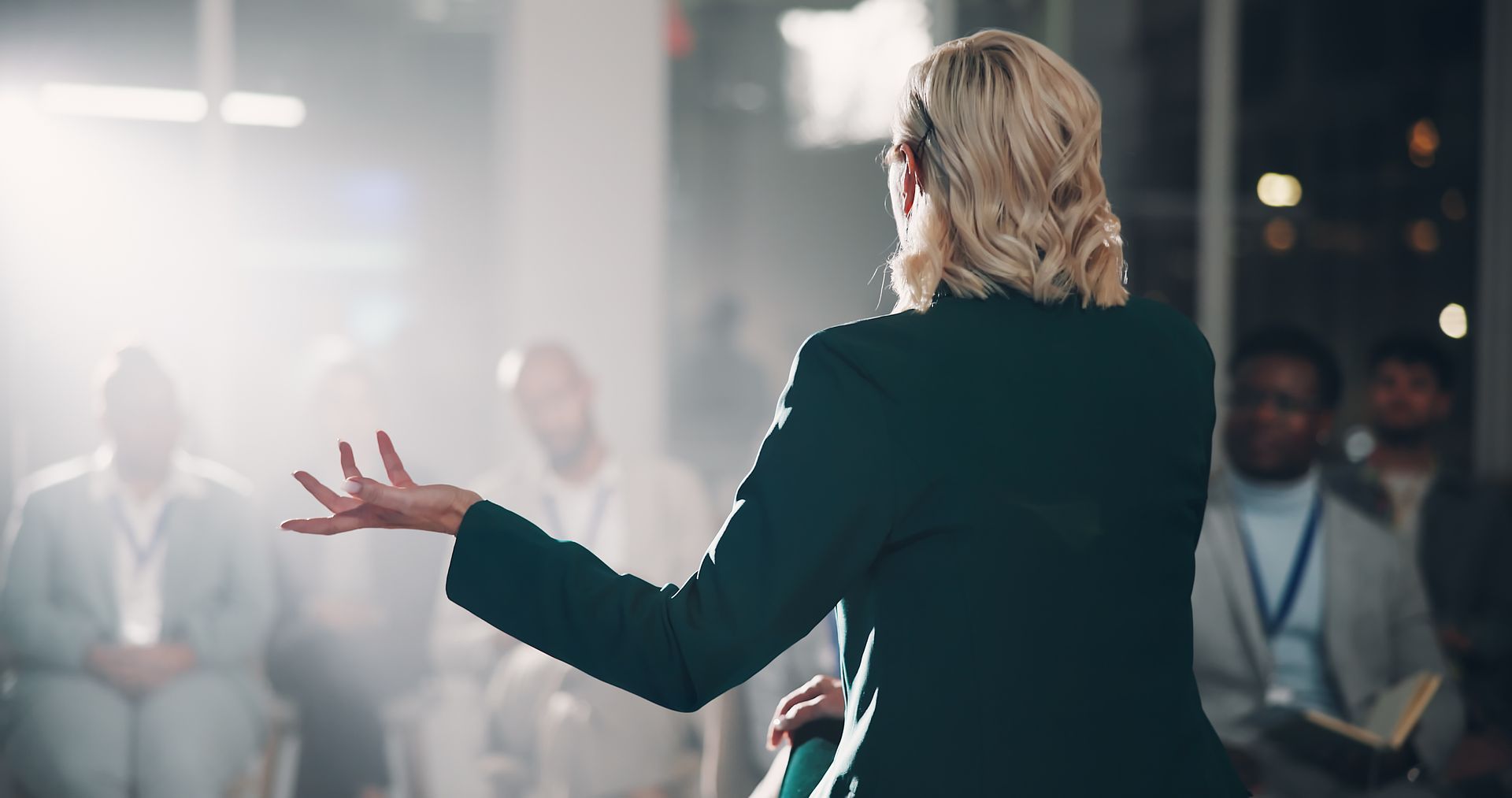 A person in a dark suit stands with their back to the camera, gesturing while addressing a blurred audience in a room. A person in a dark suit stands with their back to the camera, gesturing while addressing a blurred audience in a room.