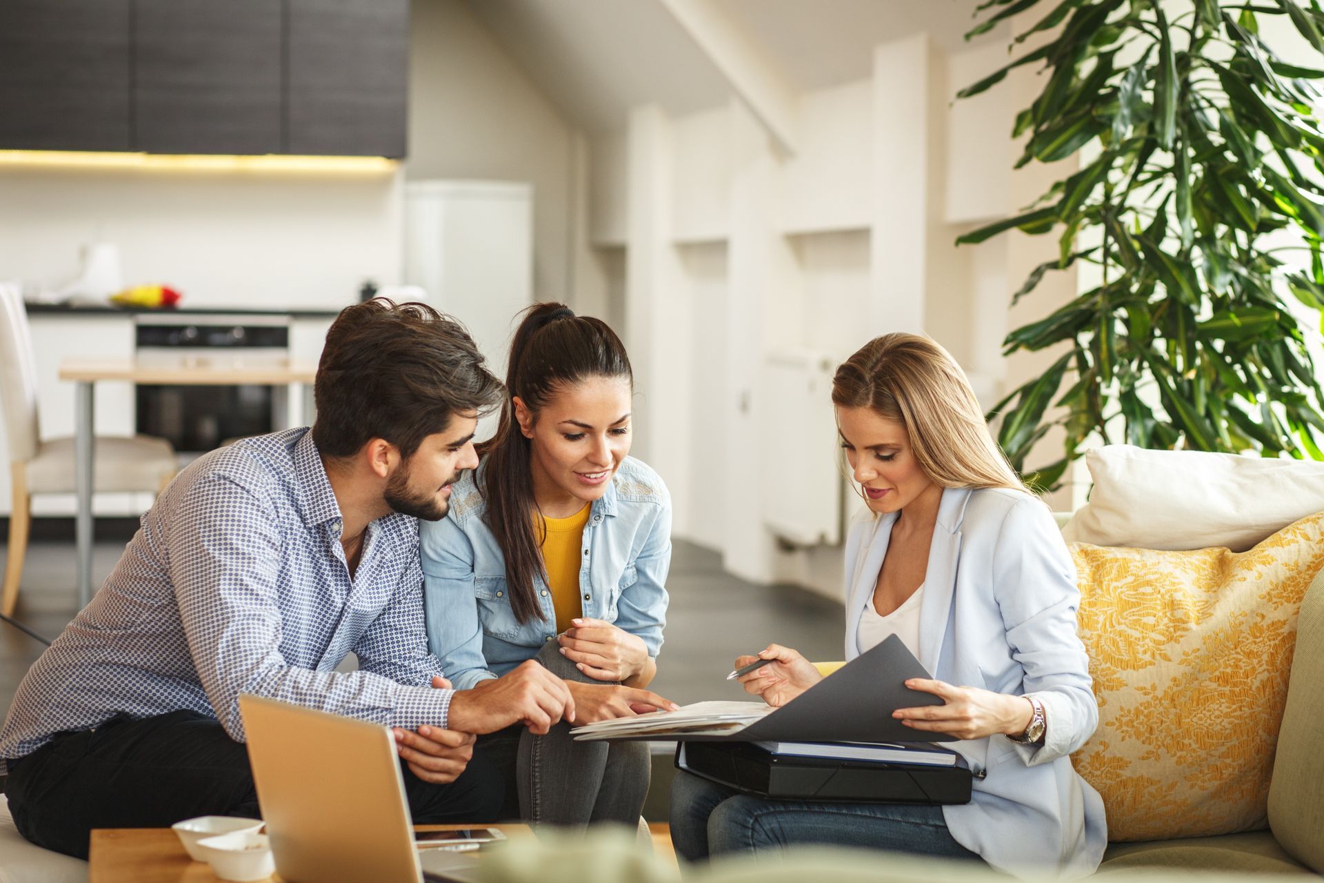 A professional consultant reviews documents with a couple in a brightly lit, modern living room.