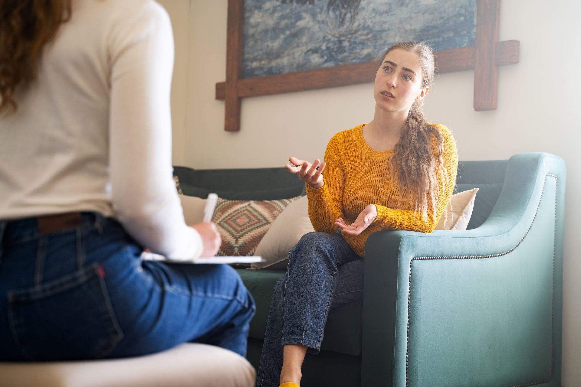 A person in a yellow sweater sits on a sofa, gesturing while speaking to a therapist who is taking notes. A person in a yellow sweater sits on a sofa, gesturing while speaking to a therapist who is taking notes.