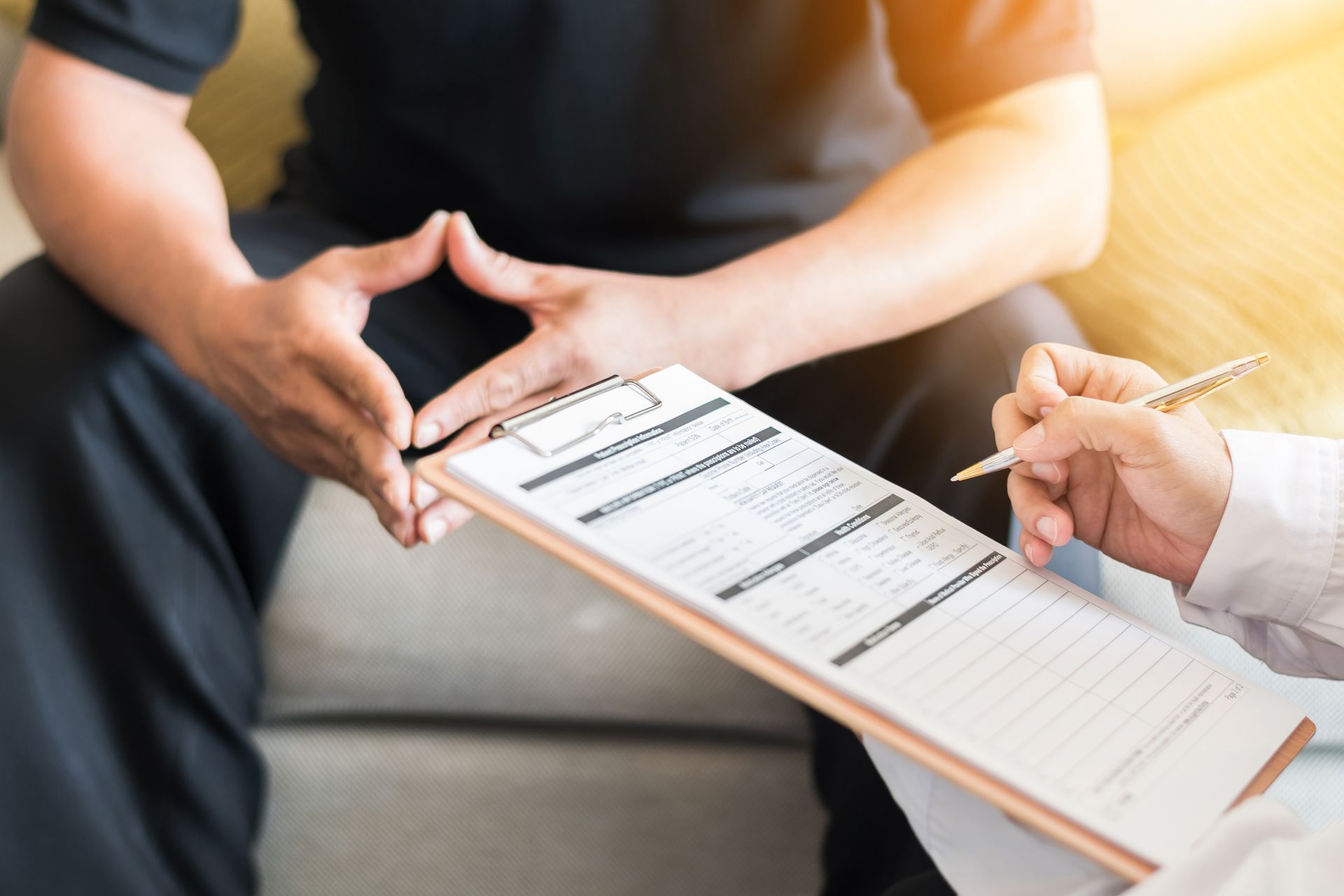 A person’s hand holds a pen to a clipboard while another person sits opposite them during a consultation.