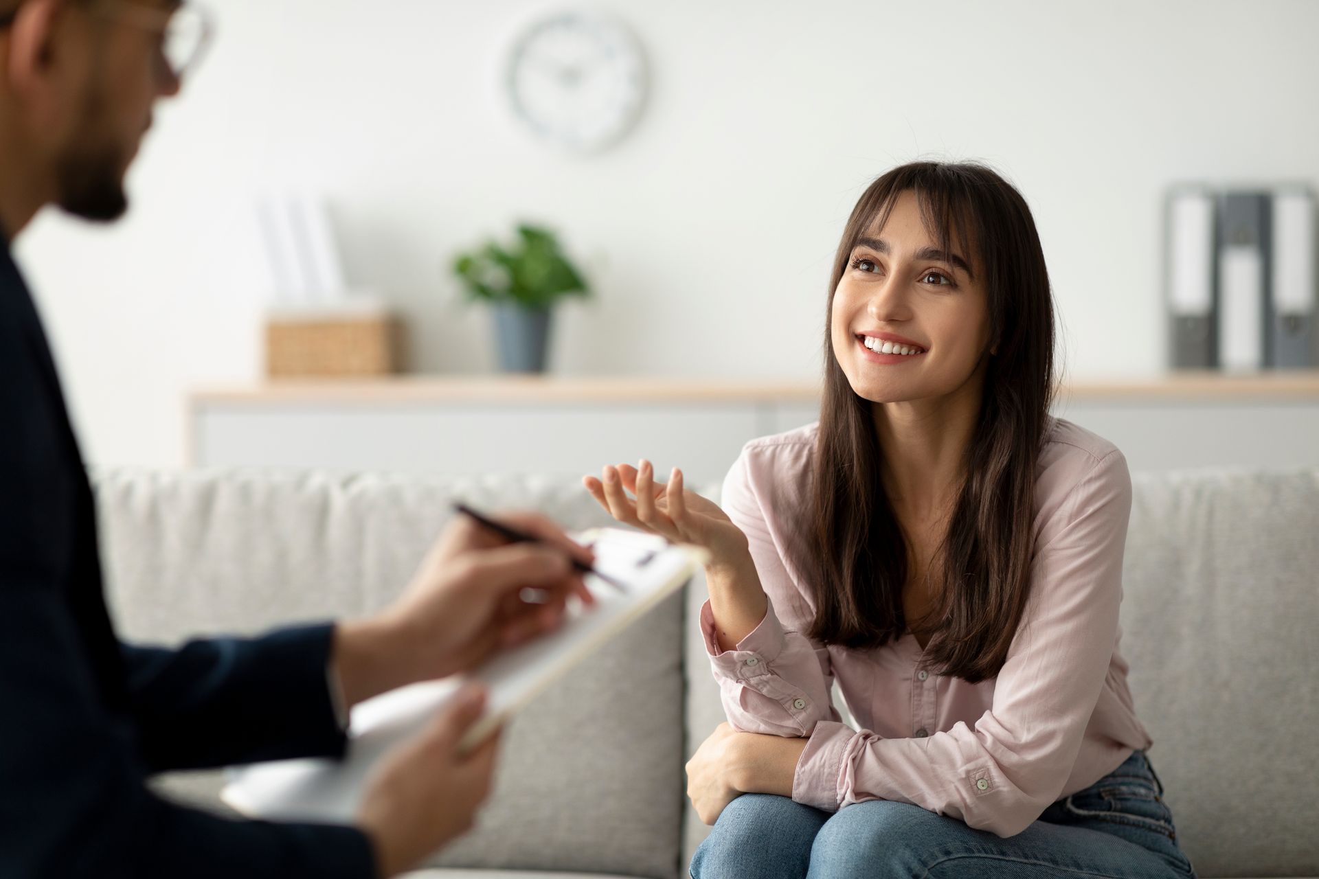 A person sits on a sofa, smiling and speaking to a professional who is taking notes on a clipboard in a bright office.