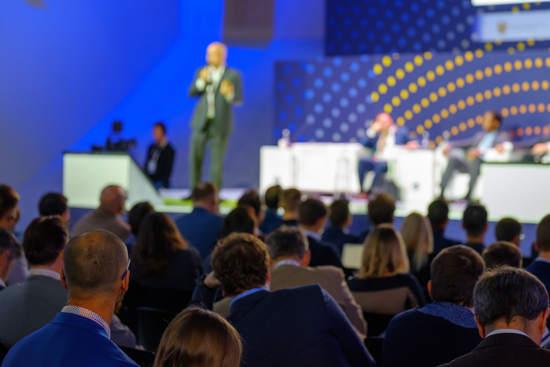 A speaker stands on a stage before an audience at an indoor conference with blue lighting and geometric background panels. A speaker stands on a stage before an audience at an indoor conference with blue lighting and geometric background panels.