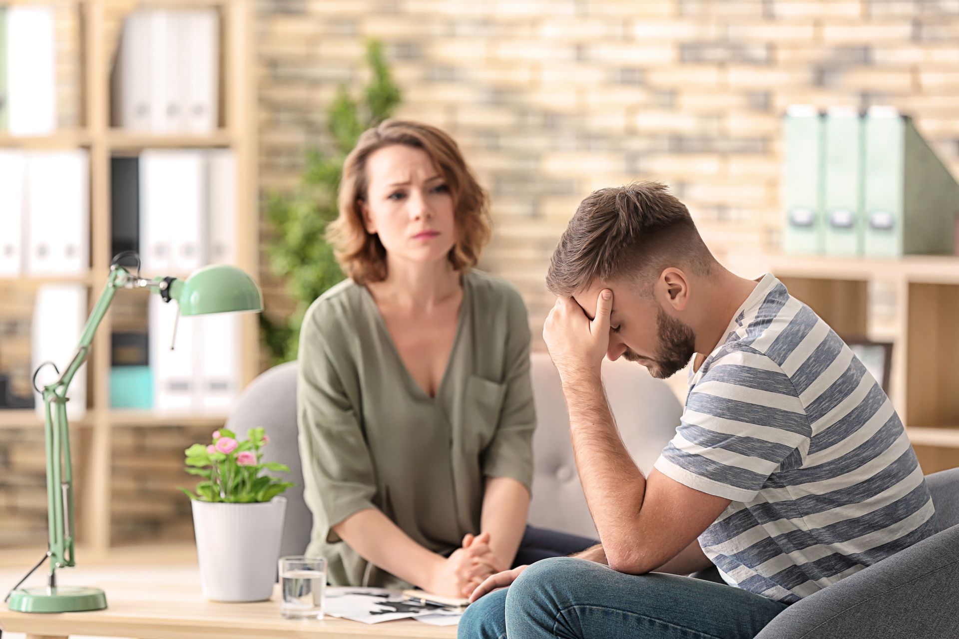 A woman looks concerned while sitting opposite a distressed man with his head in his hands in an office setting. A woman looks concerned while sitting opposite a distressed man with his head in his hands in an office setting.