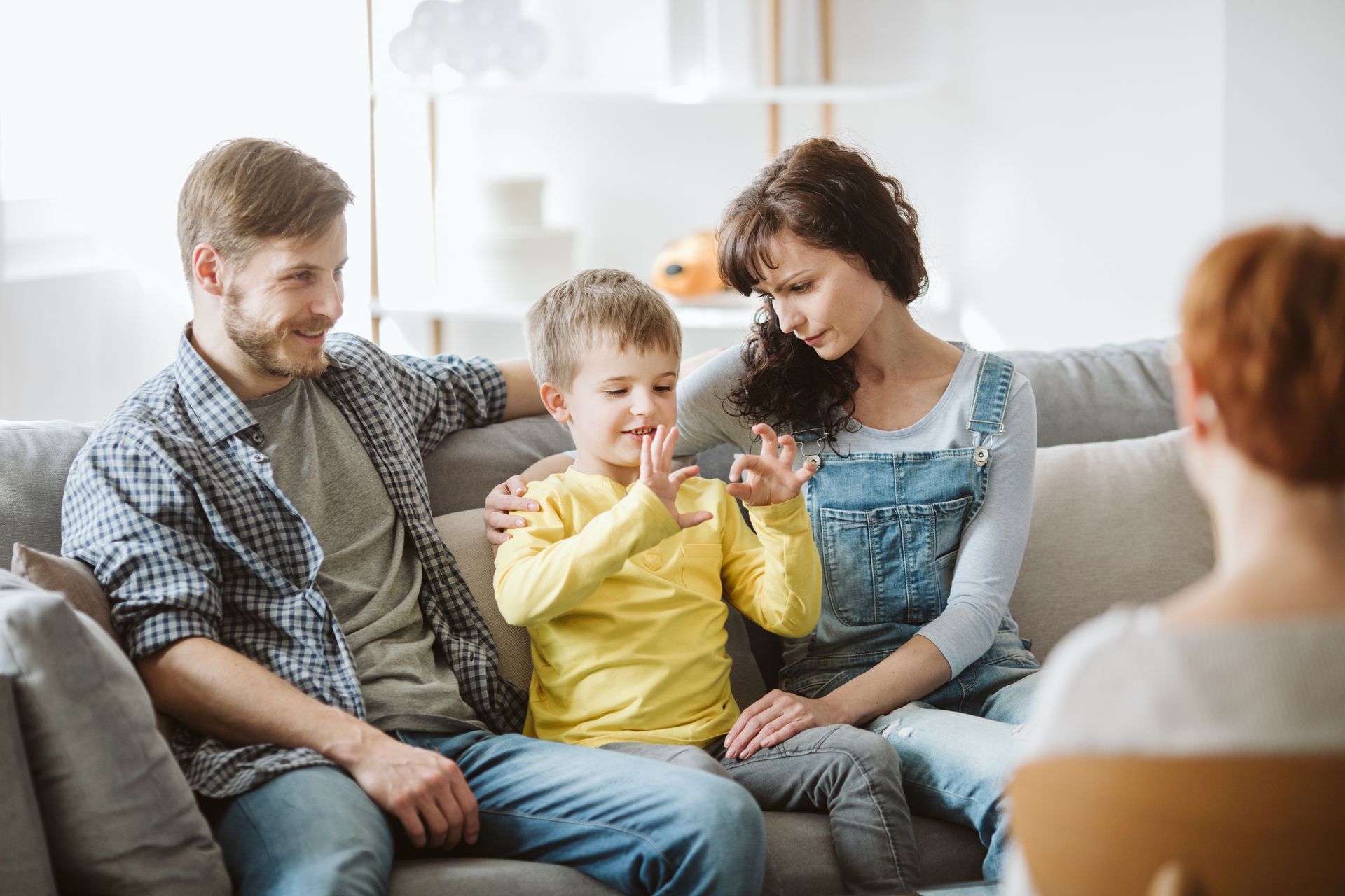 A family sits on a couch during a therapy session, with a young child making hand gestures while talking to a therapist. A family sits on a couch during a therapy session, with a young child making hand gestures while talking to a therapist.