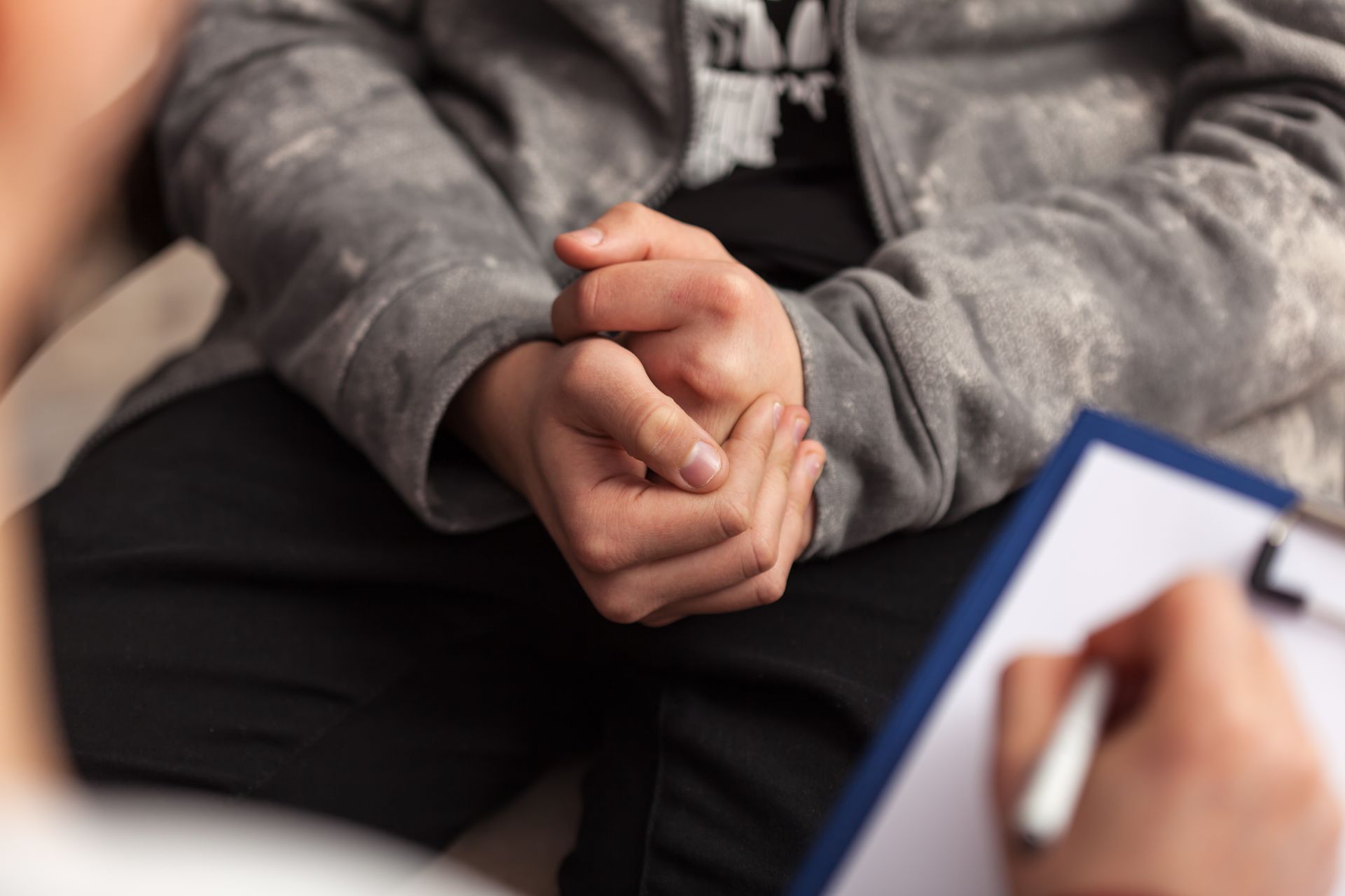 A person with clasped hands sits before an interviewer, who holds a clipboard and pen.