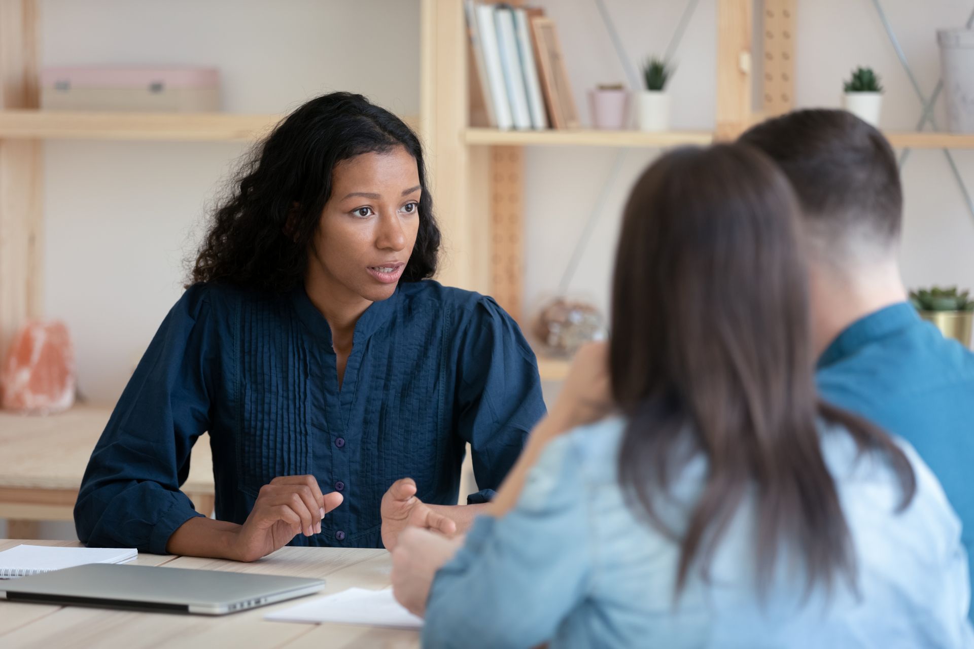A consultant speaks to a couple across a table in a professional office setting with bookshelves in the background.