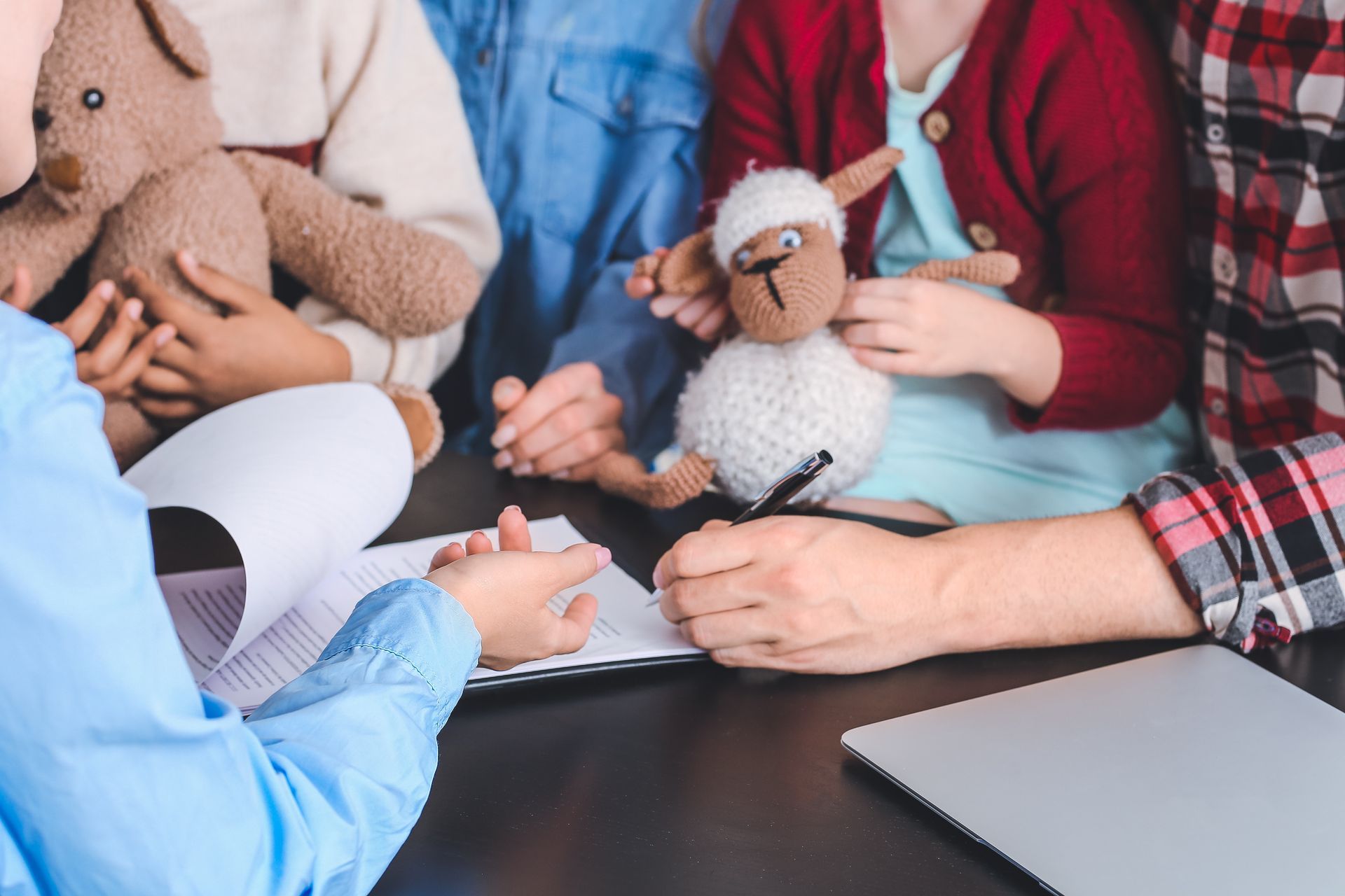 A person with a document interacts with others holding a teddy bear and a stuffed sheep at a table with a laptop.