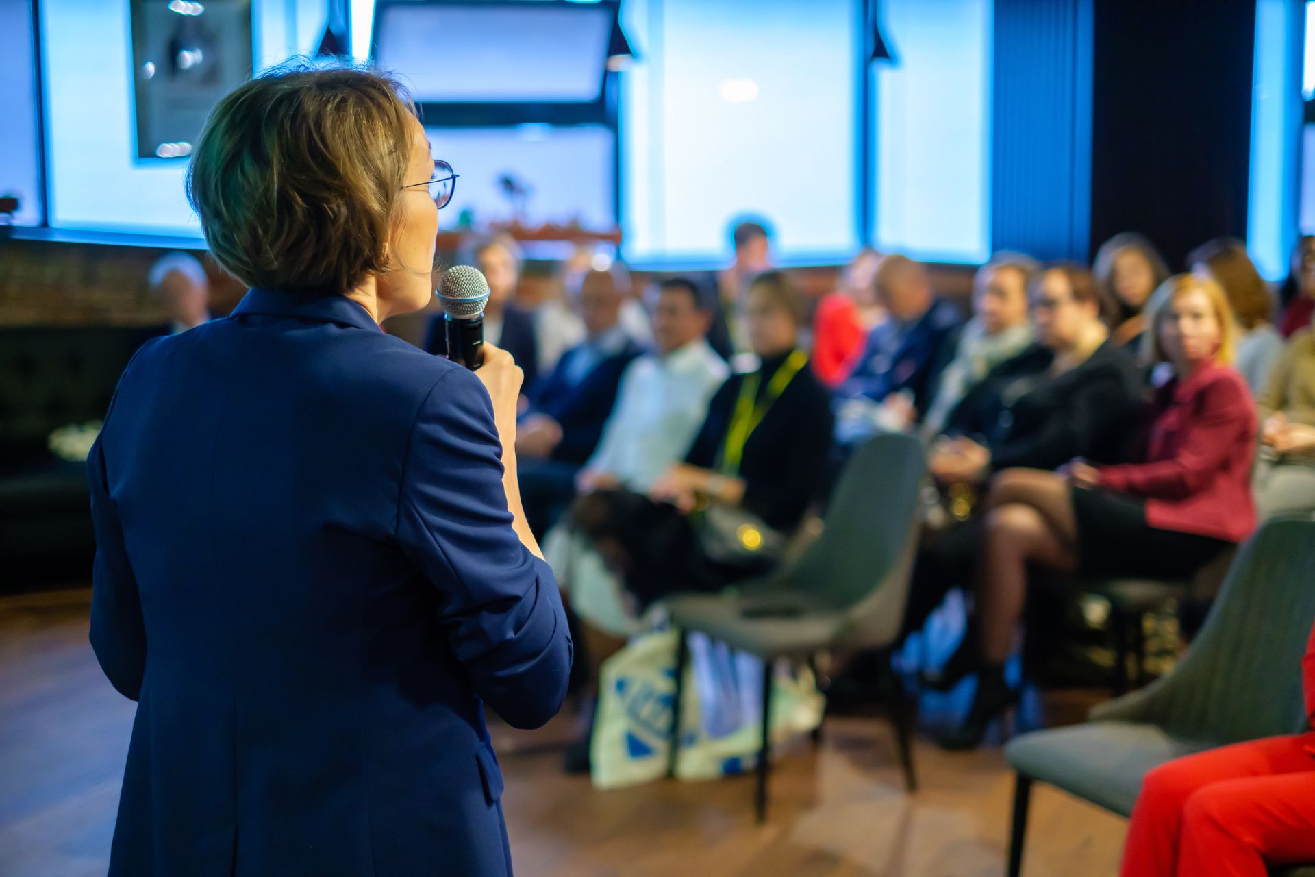 A speaker in a blue blazer stands before an audience in a room with blue-lit windows, holding a microphone. A speaker in a blue blazer stands before an audience in a room with blue-lit windows, holding a microphone.