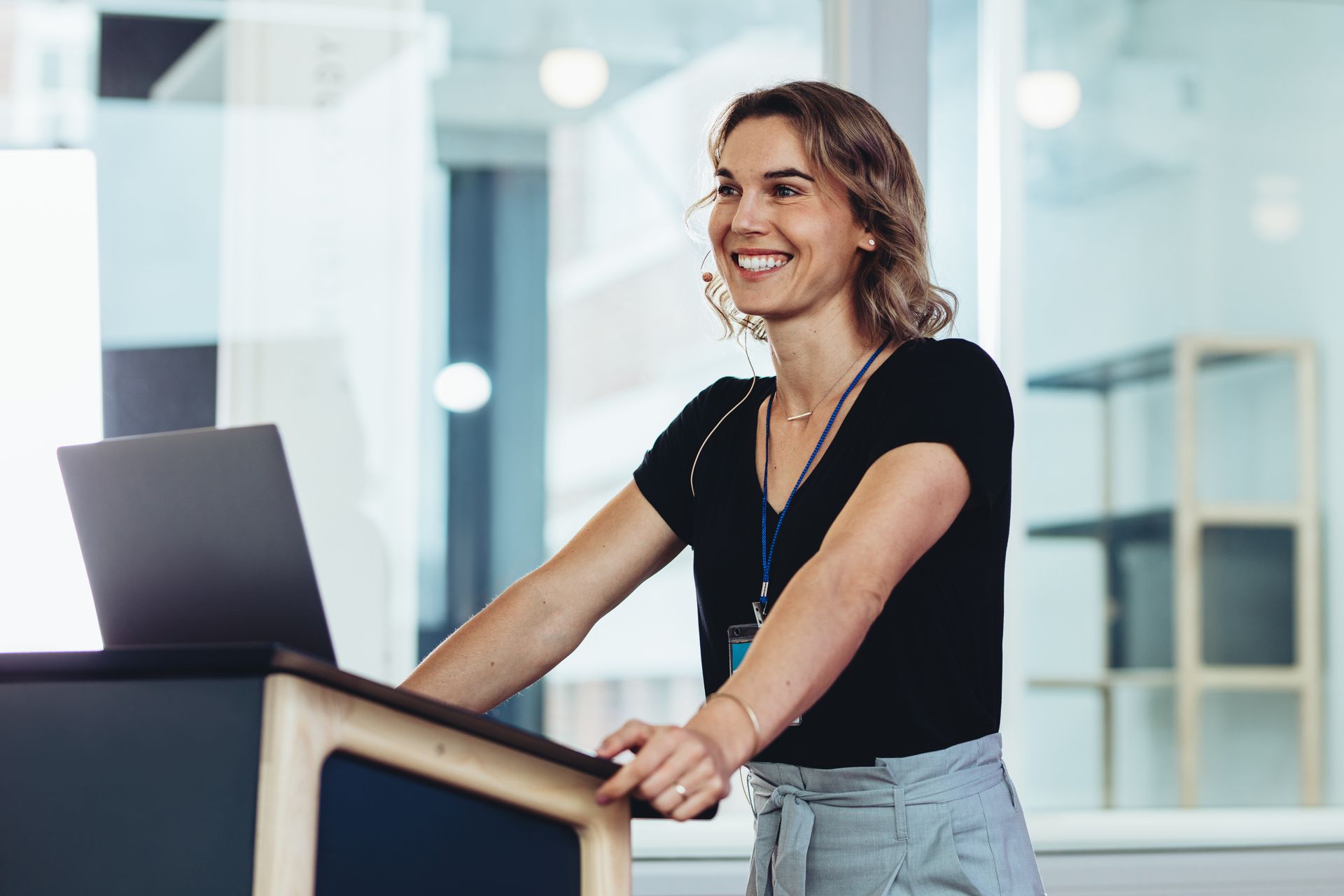 A smiling person stands at a podium in a brightly lit office, resting their hands on a laptop.