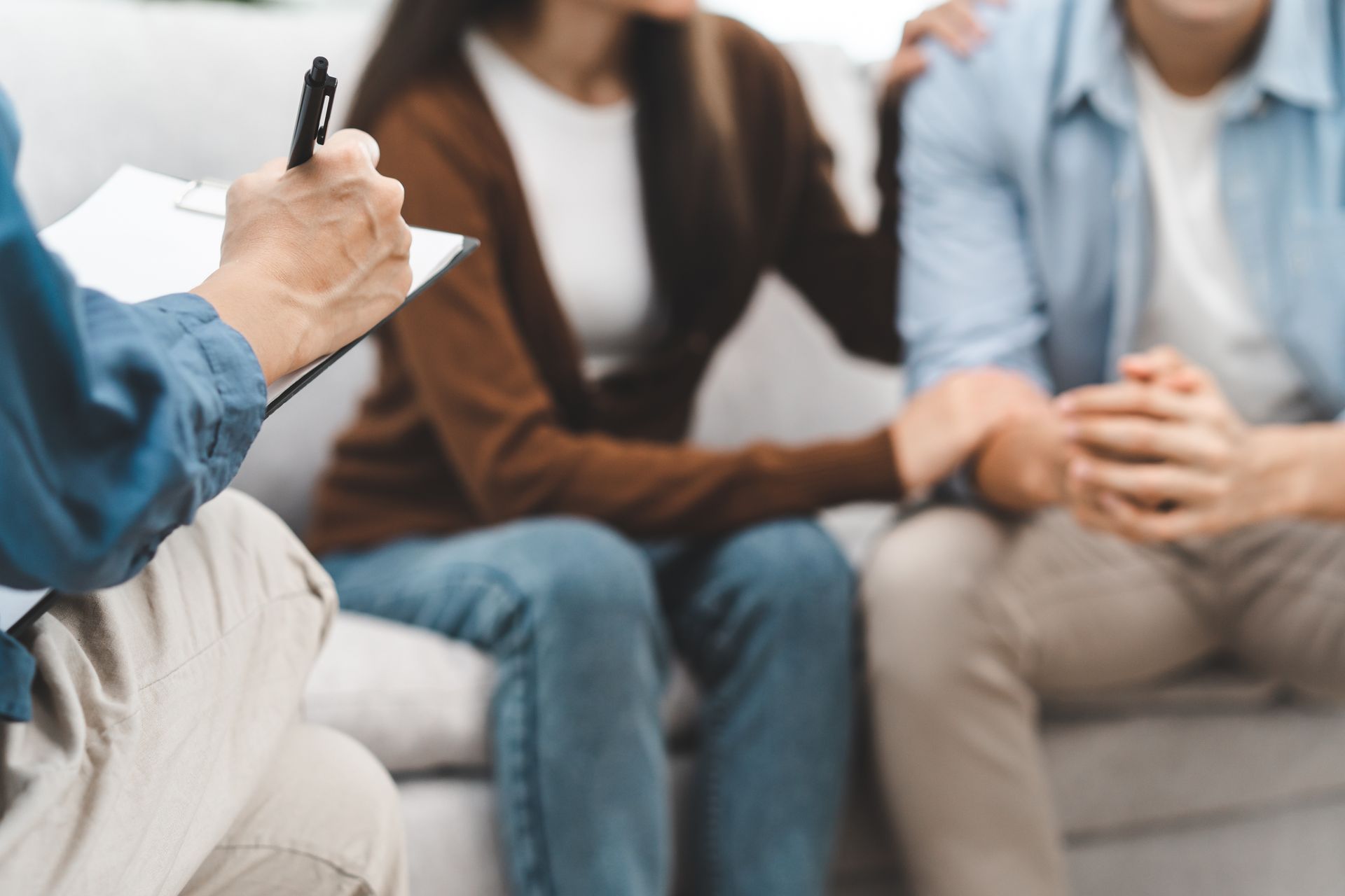 A person with a notepad and pen sits across from a pair of people sitting on a couch, suggesting a counseling session.
