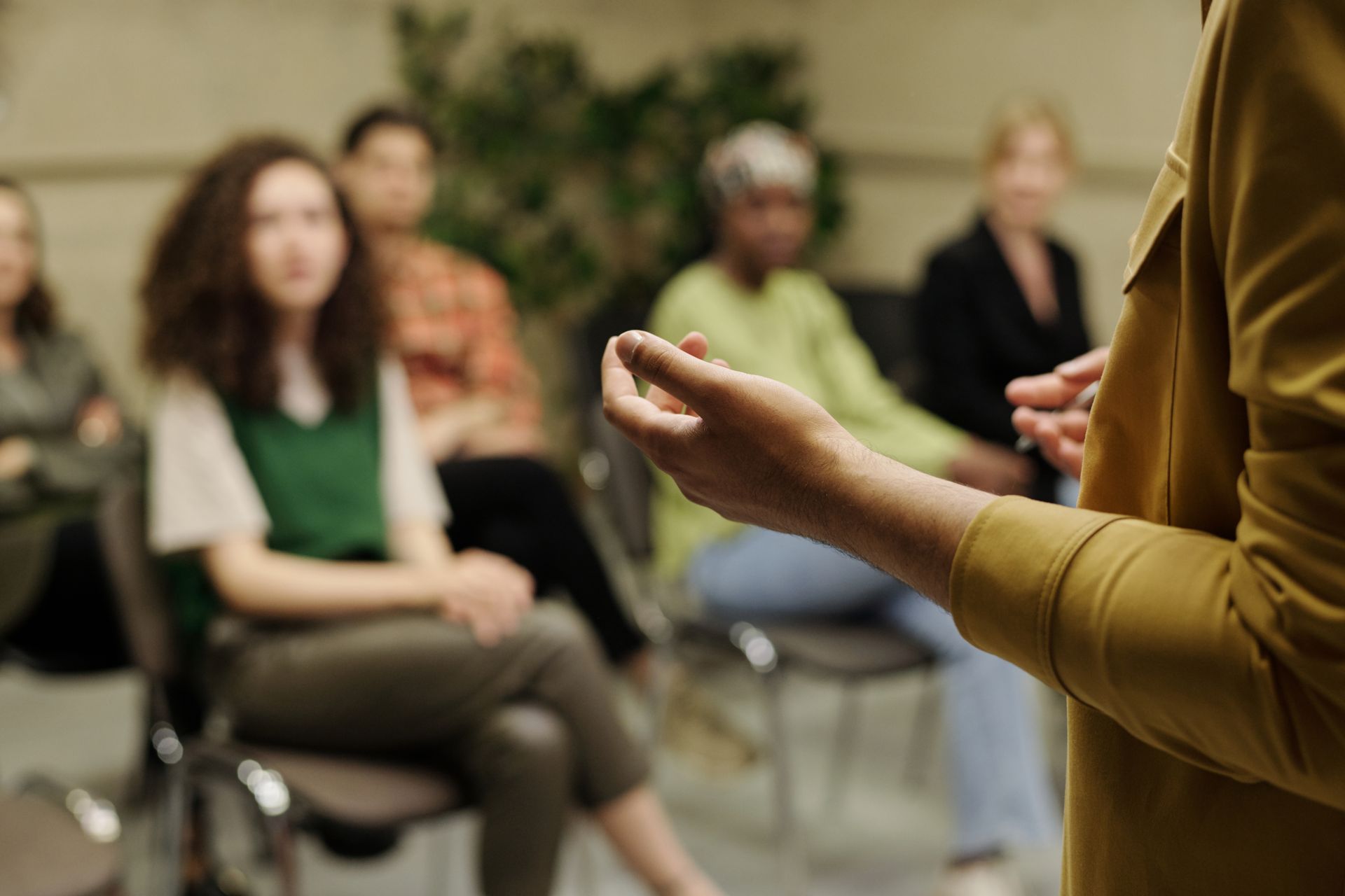 A person in a yellow jacket gestures while speaking to a group of people seated in a workshop or meeting setting.