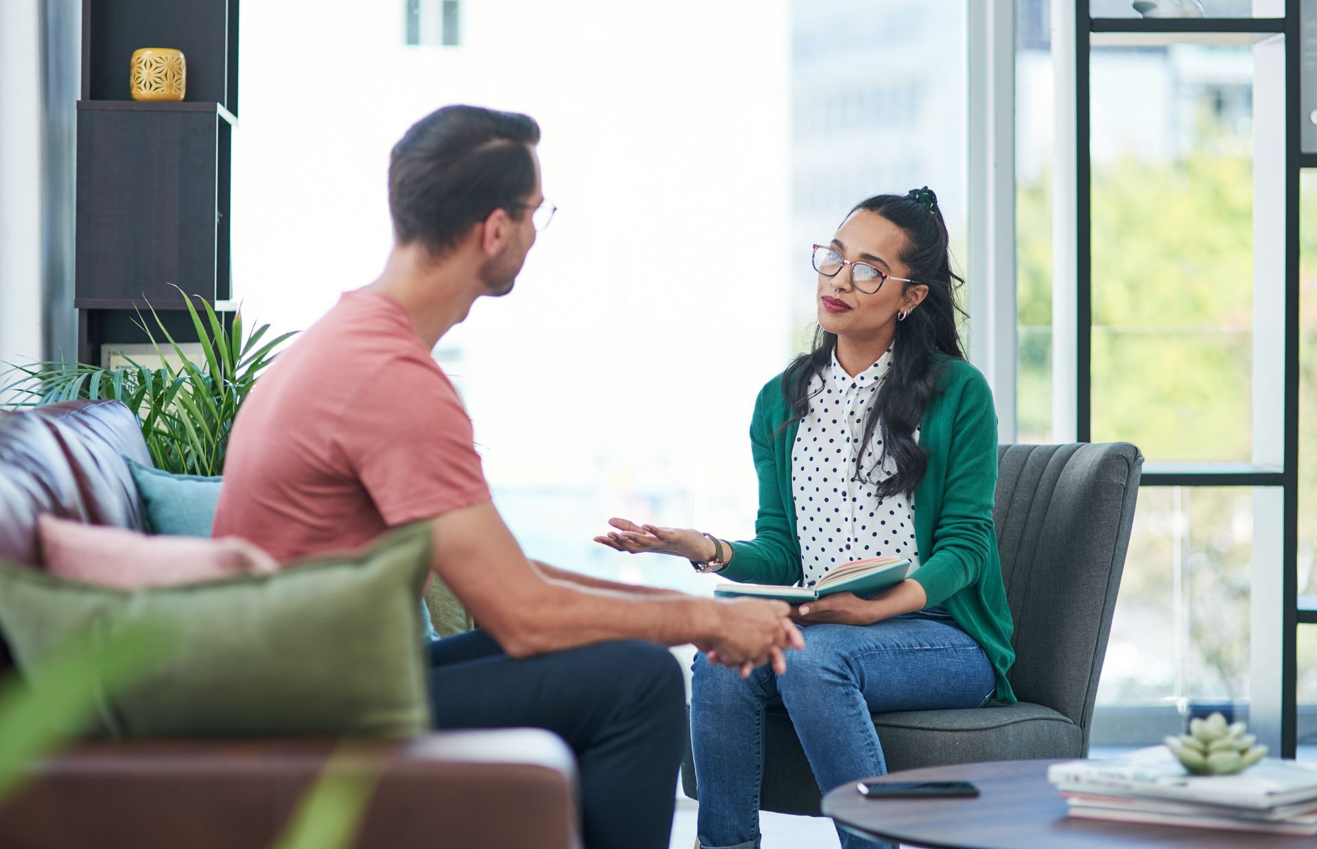 A therapist in a green cardigan holds a notebook while talking to a client sitting on a sofa in a bright, modern office. A therapist in a green cardigan holds a notebook while talking to a client sitting on a sofa in a bright, modern office.