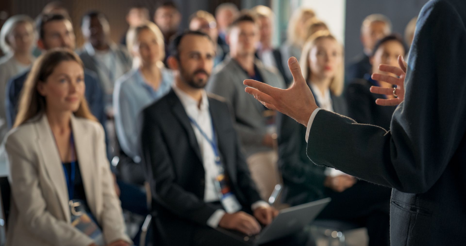 A speaker gesturing to an engaged audience in a professional conference room setting. A speaker gesturing to an engaged audience in a professional conference room setting.