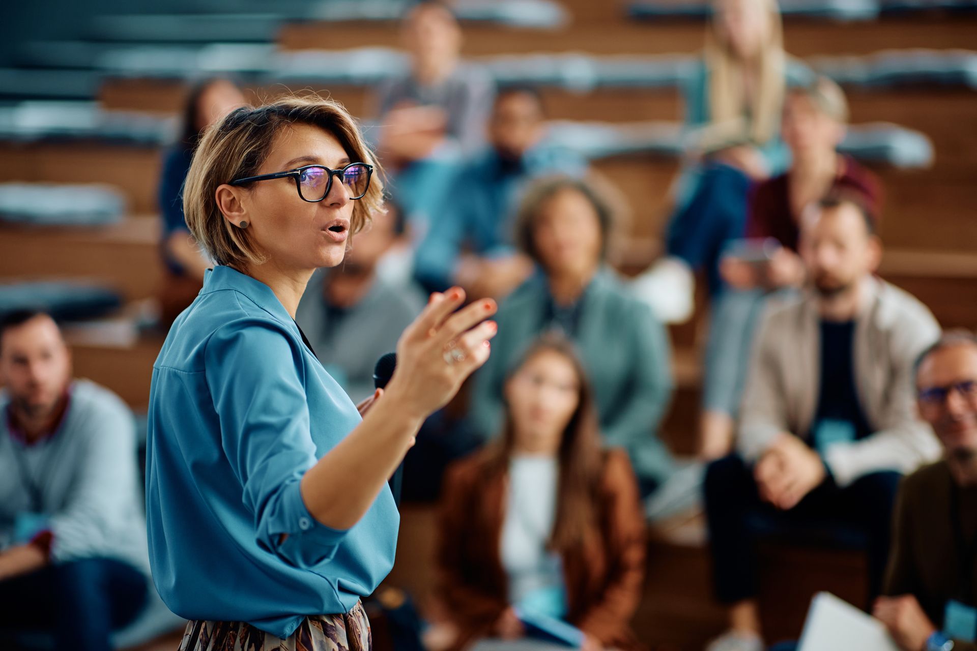 A speaker in a blue shirt gestures while presenting to an audience seated in a tiered auditorium. A speaker in a blue shirt gestures while presenting to an audience seated in a tiered auditorium.