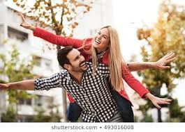 A person in a red jacket gives someone in a checkered shirt a piggyback ride, both with arms outstretched outdoors.
