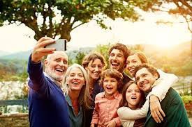 A happy, multi-generational family outdoors, smiling while taking a selfie together under a large tree at sunset.
