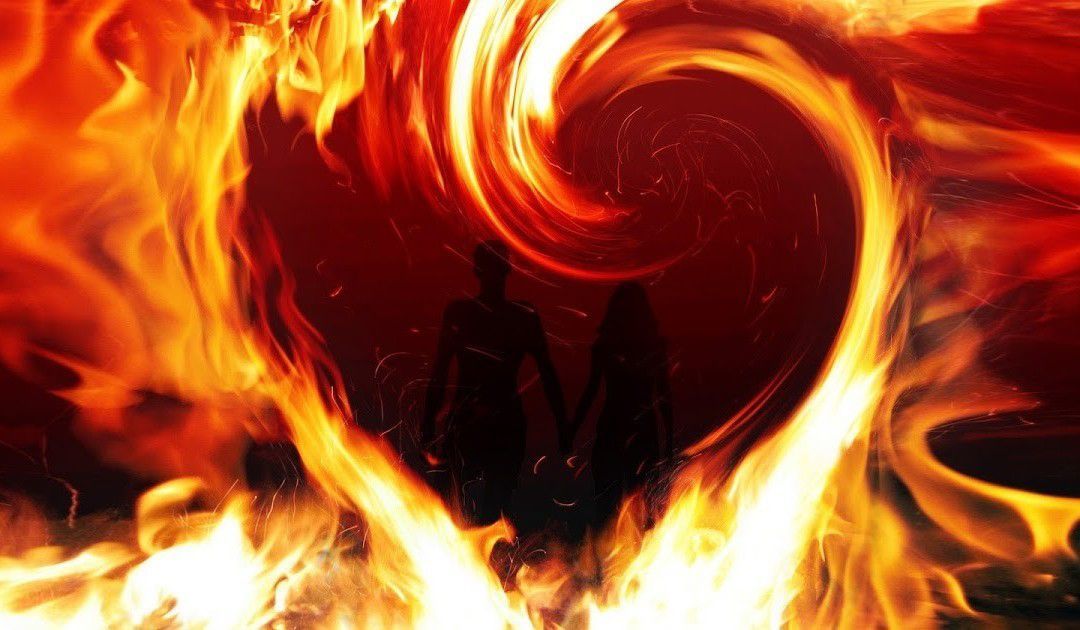 A silhouetted couple stands hand-in-hand inside a glowing, heart-shaped vortex of bright orange and yellow flames.