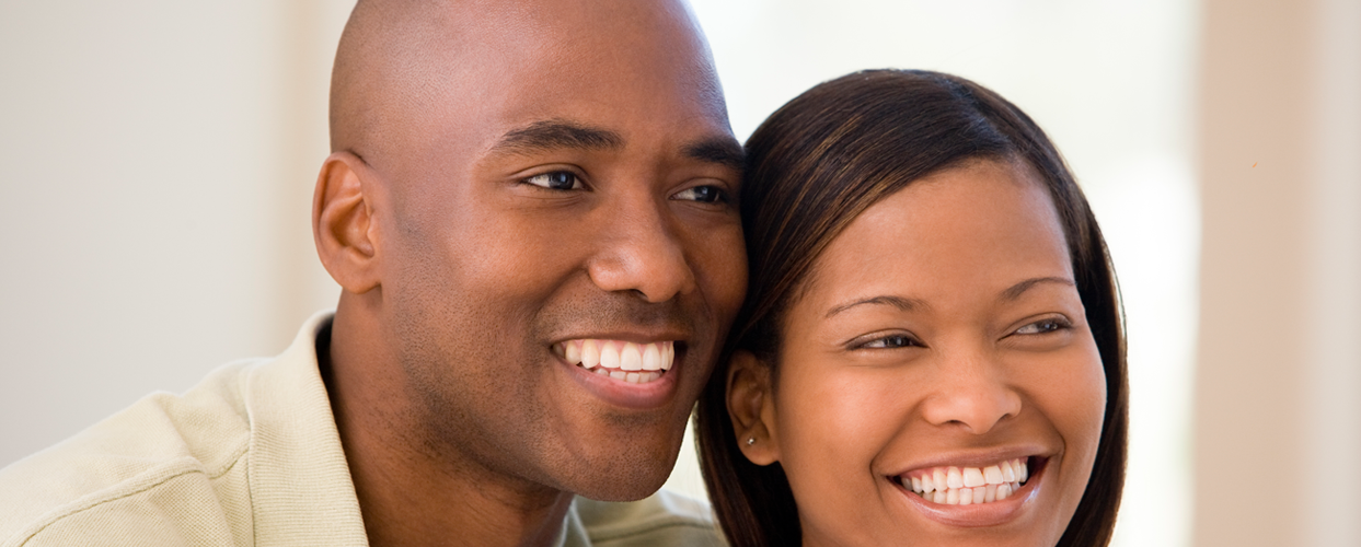 Smiling couple close together, indoors. Man with shaved head, woman with dark hair.