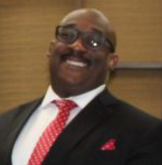 Man smiling in a suit and tie; red patterned tie, black suit, and glasses.