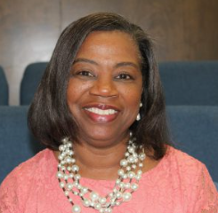 Woman with short dark hair, smiling, wearing a pink top and layered pearl necklace.