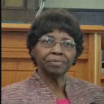 Woman wearing glasses, pink top, and patterned blazer in a wood-paneled room.