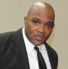 Man in a black suit and tie, looking towards the camera against a neutral background.