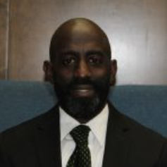Man in a suit with a green tie and black beard, sitting in front of a blue background.