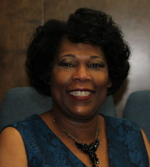 Woman smiling, wearing a dark blue dress and necklace, seated indoors.