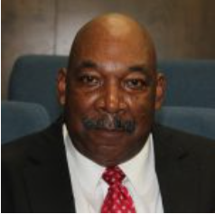 Man in suit and tie with a mustache, seated in front of a blue background.
