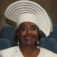 Woman wearing a white hat, smiling in a room with blue seats.