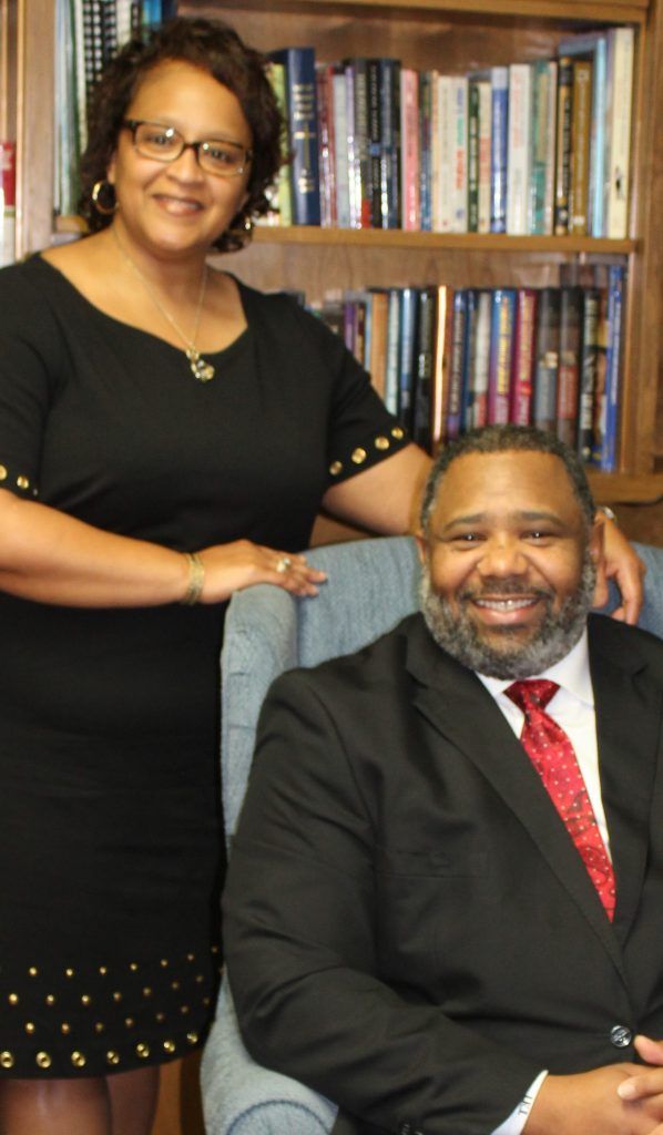 Woman stands behind a man seated in front of a bookshelf. Both are smiling. He wears a suit. She wears a black dress.