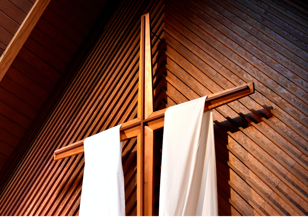 Wooden cross with white draped cloth, set against wood-paneled wall.