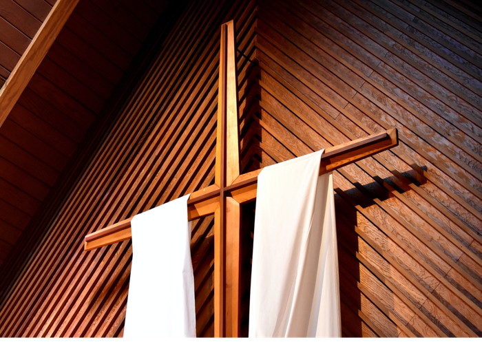 Wooden cross with white draped cloth, set against wood-paneled wall.