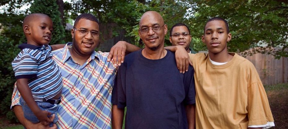 Five people of color smiling together, posing outside with a wooden fence and greenery in the background.
