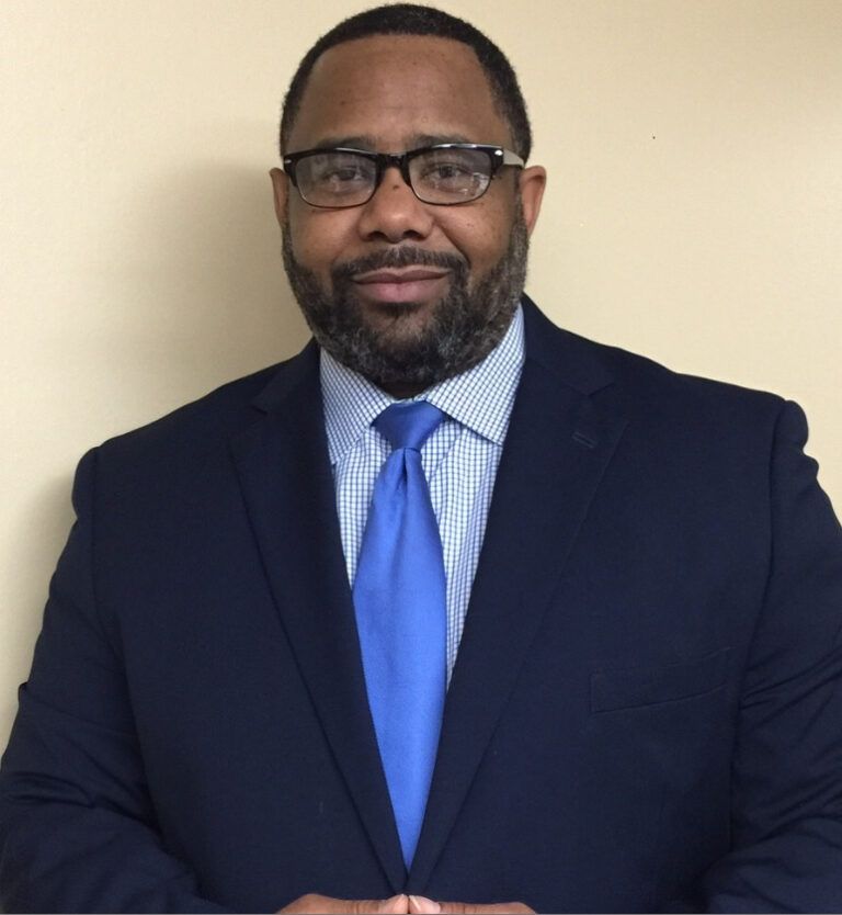Man in a navy blue suit, blue tie, and glasses, smiling, set against a light background.