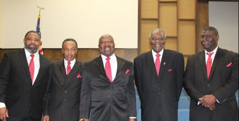 Five men in black suits and red ties pose together indoors.