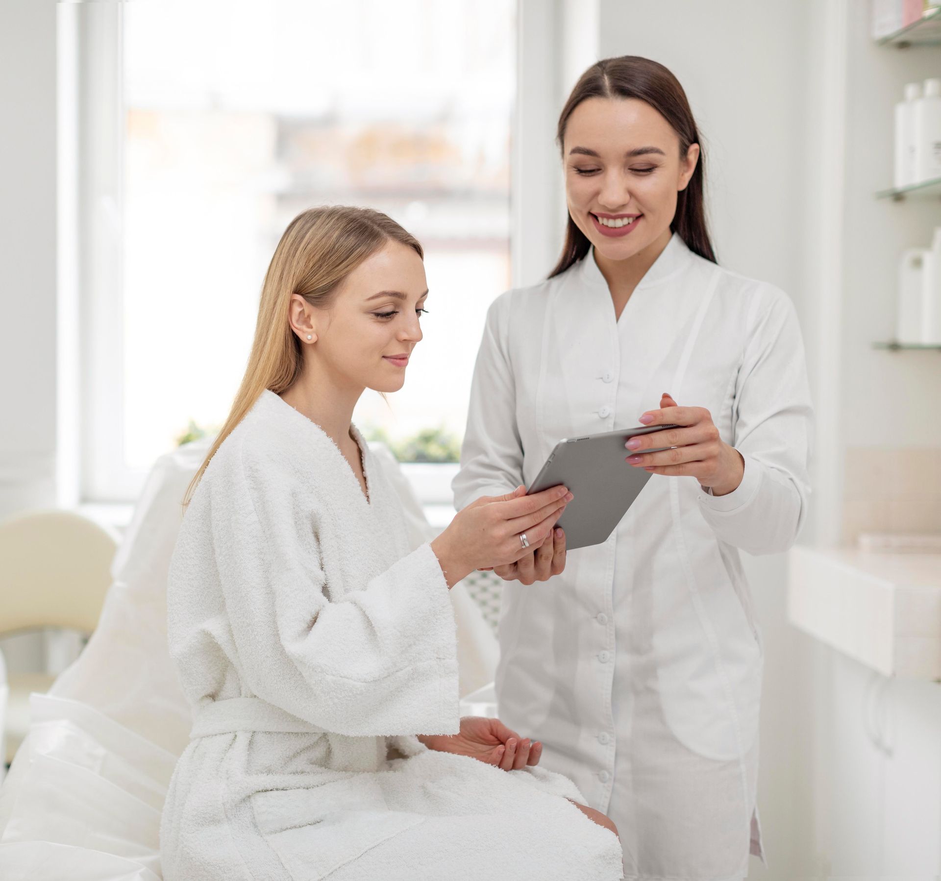 Woman in robe consulting with a healthcare professional in a clinic, viewing tablet.