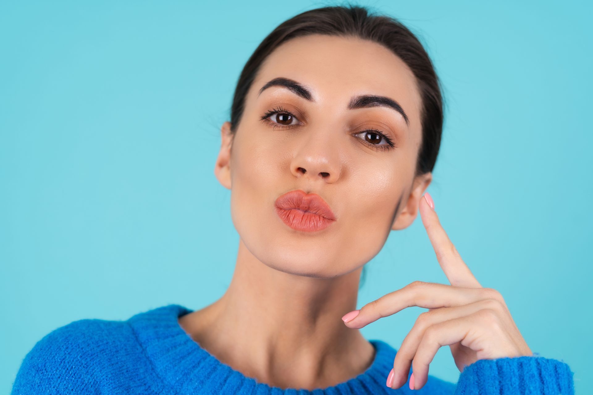 A woman in a blue sweater is blowing a kiss and touching her ear.